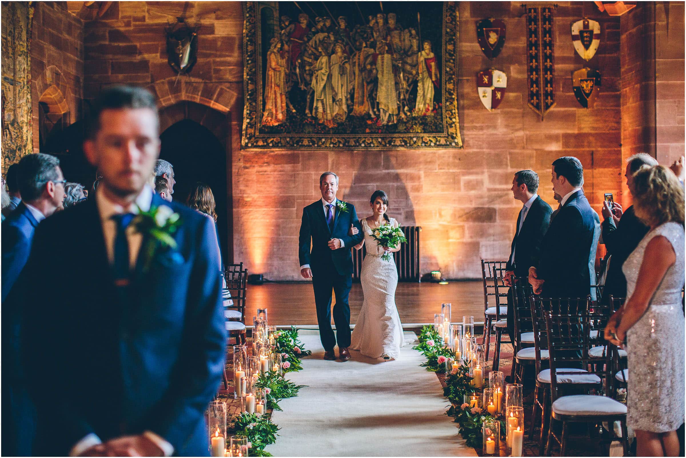 A bride walks down the candlelit aisle at Peckforton Castle while the groom waits at the altar. Guests turn to face her