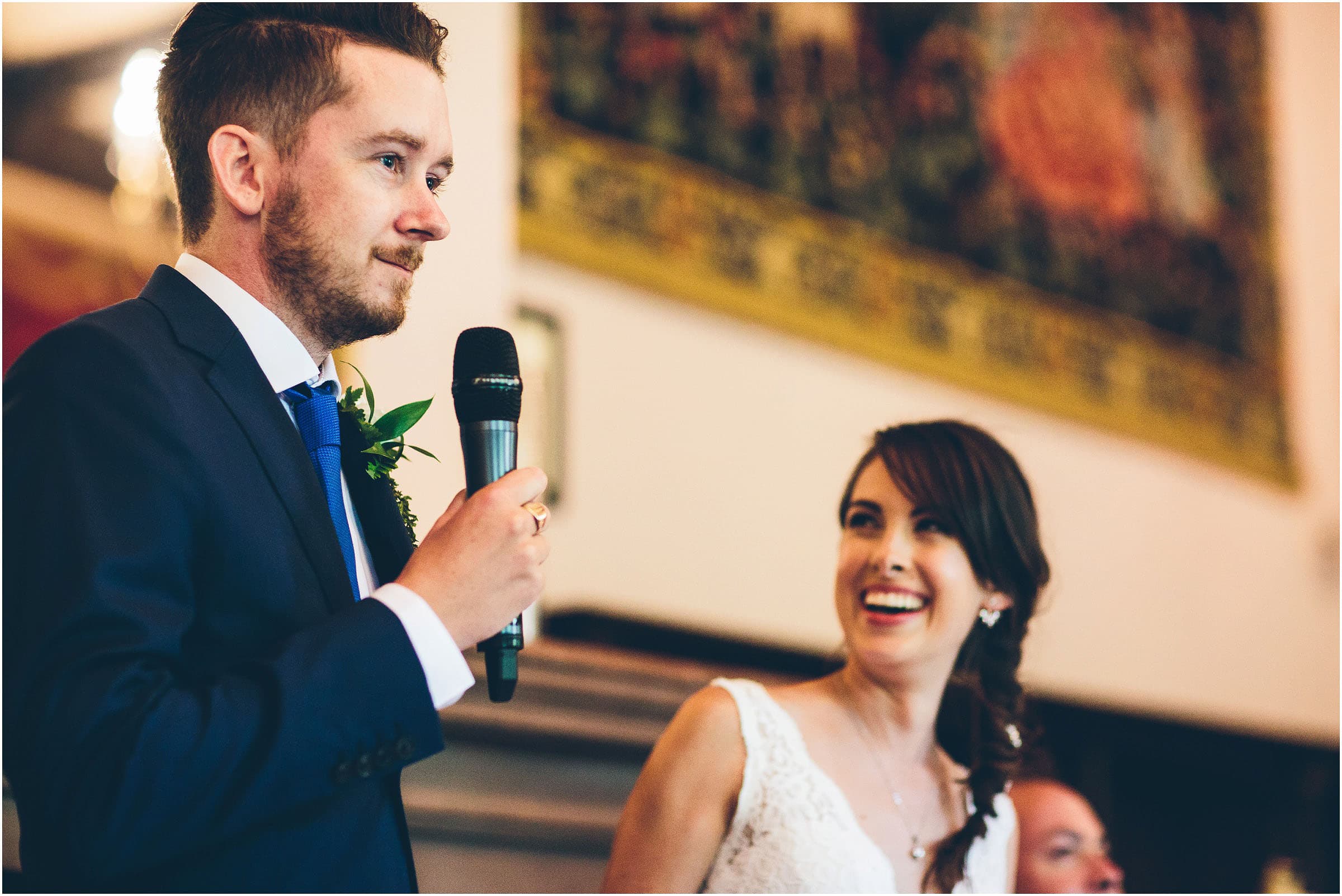 A groom giving his speech in front of a large tapestry at Peckforton Castle while his bride laughs in the background.