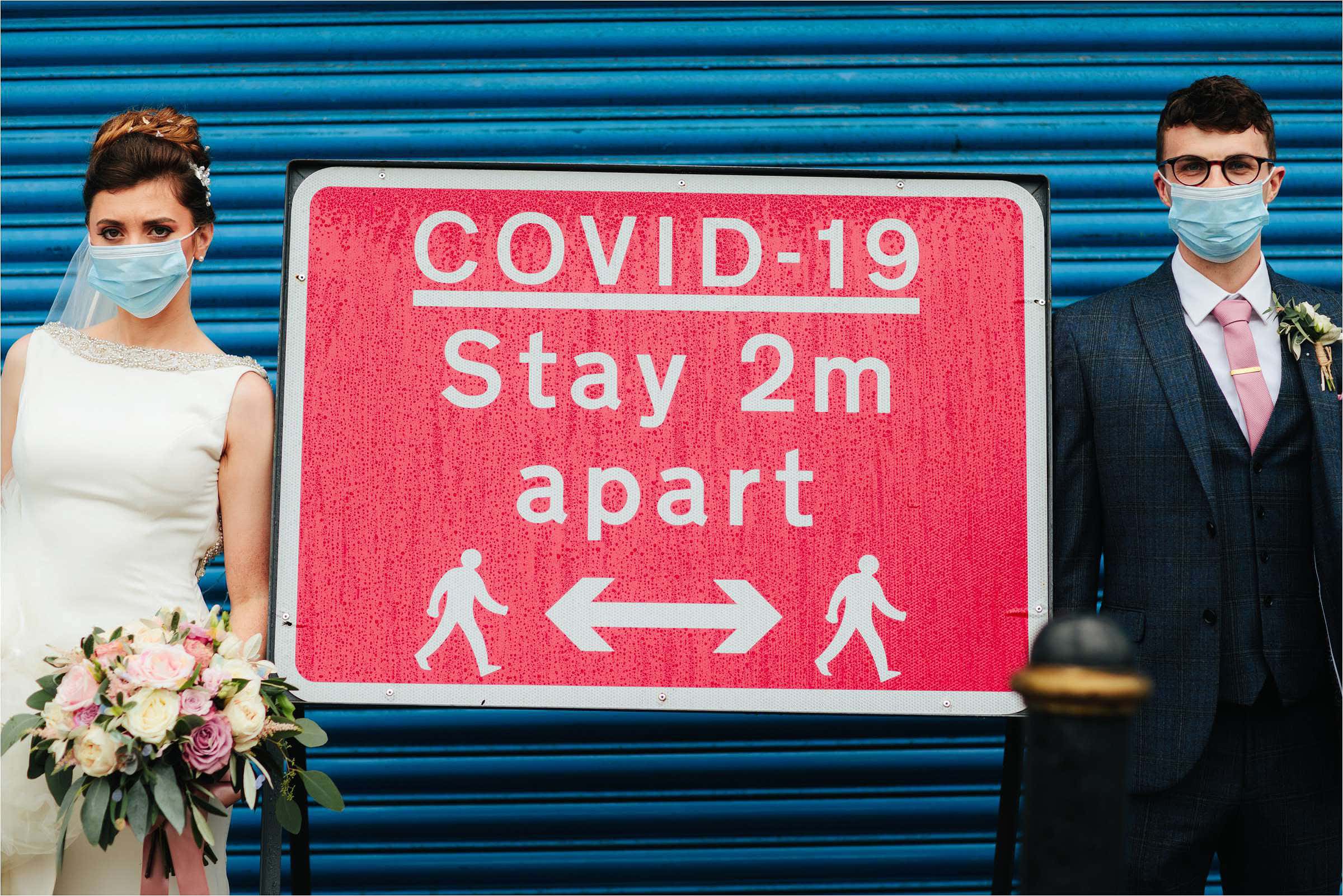 bride and groom standing against blue backdrop either side of covid-19 stay 2m part red sign
